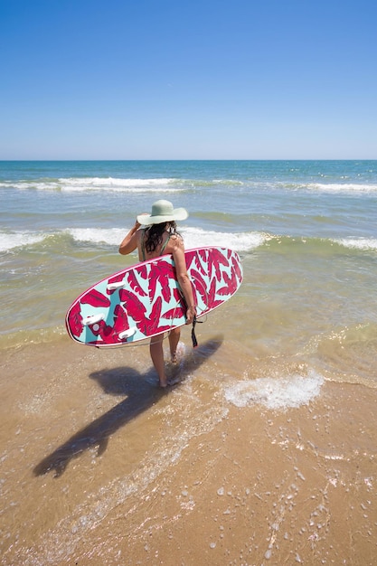 Aantrekkelijke gelukkige vrouw op middelbare leeftijd die een hoedzonnebril draagt en een surfplank op het strand draagt