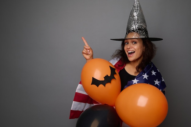 Aantrekkelijke gemengd ras vrouw in tovenaarshoed, gewikkeld in Amerikaanse vlag, houdt kleurrijke oranje luchtballen vast, glimlacht brede glimlach kijkend naar camera, wijst op kopieerruimte op grijze achtergrond. Halloween-concept