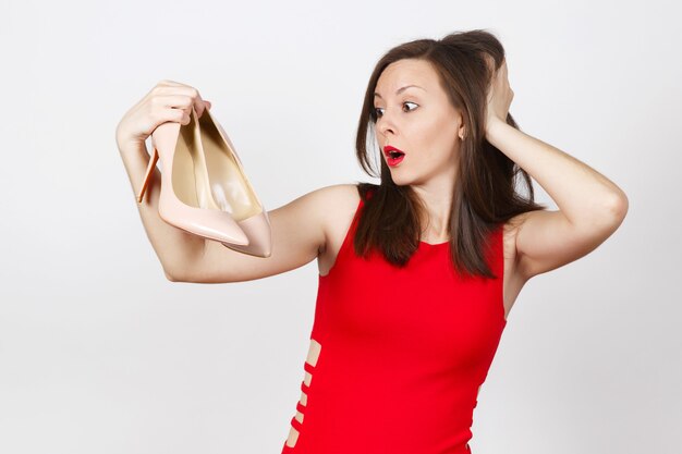 Aantrekkelijke glamour Kaukasische modieuze jonge peinzende verbijsterd bruin haar vrouw in rode jurk beige schoenen krijgen met rode zool van winkelen geïsoleerd op een witte achtergrond. Kopieer ruimte voor advertentie.