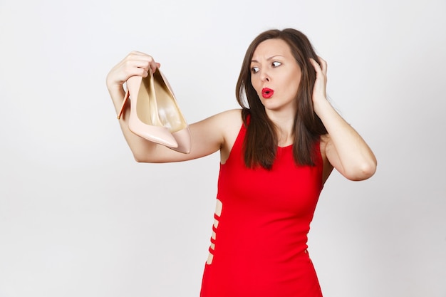 Aantrekkelijke glamour Kaukasische modieuze jonge peinzende verbijsterd bruin haar vrouw in rode jurk beige schoenen krijgen met rode zool van winkelen geïsoleerd op een witte achtergrond. Kopieer ruimte voor advertentie.