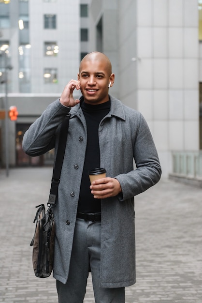 Aantrekkelijke glimlachende jonge afrikaanse man met herfstjas terwijl hij in de straat van de stad staat, naar muziek luistert terwijl hij afhaalkoffie drinkt