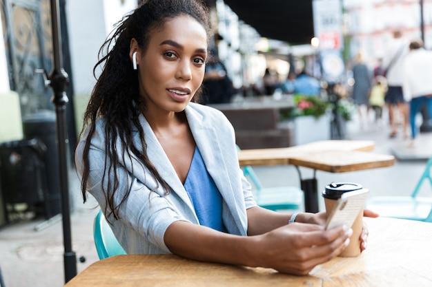 Aantrekkelijke glimlachende jonge Afrikaanse zakenvrouw die buiten aan de cafétafel zit en naar muziek luistert met draadloze oortelefoons tijdens het gebruik van de mobiele telefoon