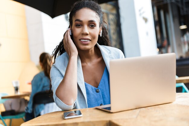 Aantrekkelijke glimlachende jonge Afrikaanse zakenvrouw die buiten aan de cafétafel zit en op een laptop werkt