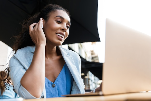 Aantrekkelijke glimlachende jonge Afrikaanse zakenvrouw die buiten aan de cafétafel zit en op een laptop werkt