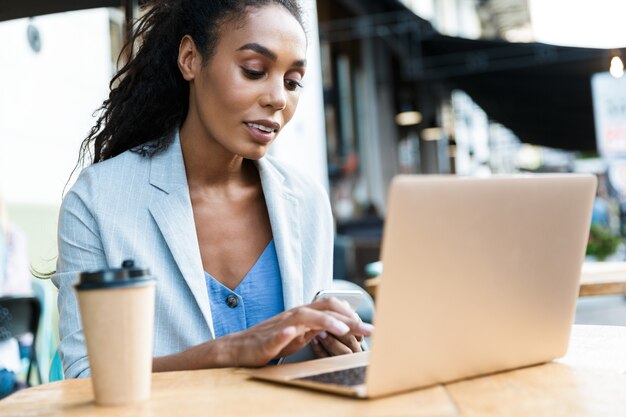 Aantrekkelijke glimlachende jonge afrikaanse zakenvrouw die buiten aan de cafétafel zit en op een laptop werkt