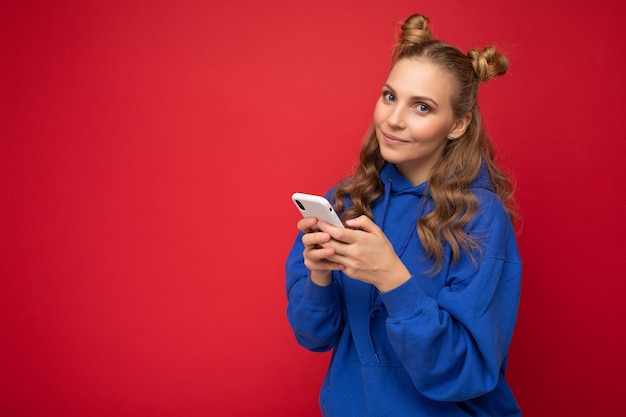 Aantrekkelijke glimlachende jonge blonde vrouw, gekleed in blauwe stijlvolle hoodie staande geïsoleerd over rode achtergrond texting