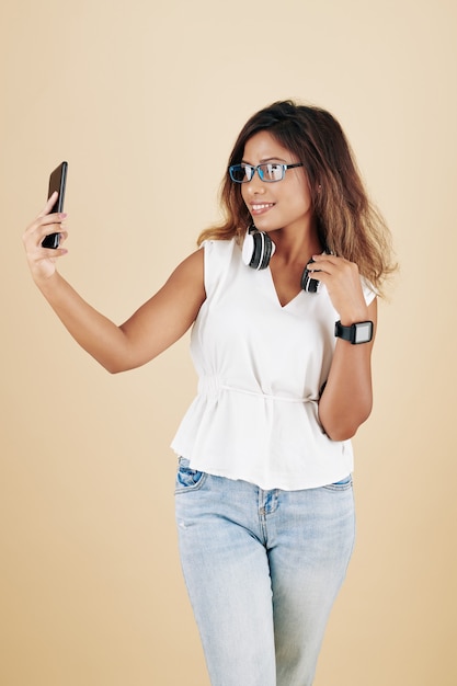 Aantrekkelijke glimlachende jonge vrouw in jeans en witte blouse die selfie op smartphone neemt