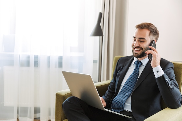 Aantrekkelijke glimlachende jonge zakenman met een pak zittend in een stoel in de hotelkamer, werkend op een laptopcomputer