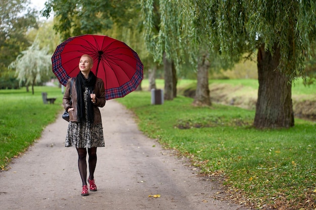 Aantrekkelijke glimlachende kortharige blonde vrouw die in het herfstpark loopt met een enorme rode paraplu