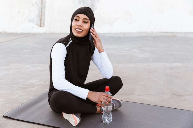 Aantrekkelijke glimlachende moslimsportvrouw die hijab buitenshuis draagt, zittend op een fitnessmat met waterfles, met behulp van mobiele telefoon