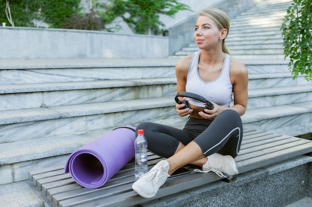 Aantrekkelijke glimlachende vrolijke sportvrouw die op een bankje rust en buiten naar muziek luistert