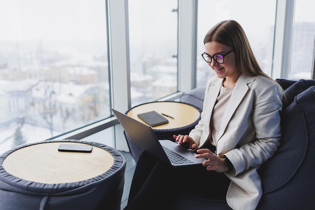 Aantrekkelijke inhoud blonde vrouw in vrijetijdskleding en bril die zich op het scherm concentreert en op laptop typt terwijl ze op een zachte poef in de lounge zit