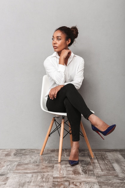 Aantrekkelijke jonge Afrikaanse bedrijfsvrouw die overhemdszitting in een geïsoleerde stoel draagt, wegkijkend