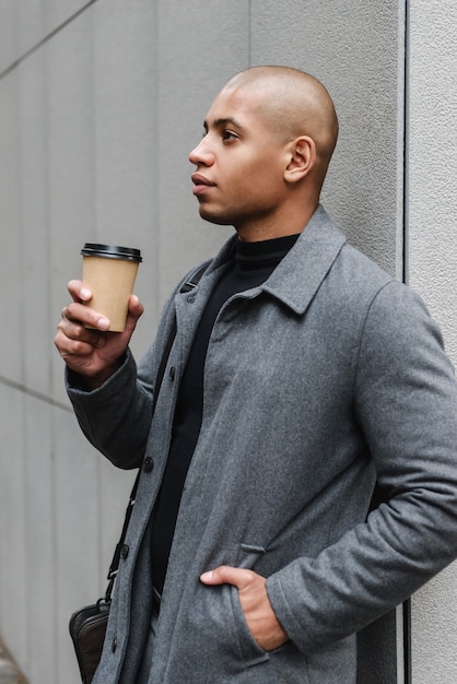 Aantrekkelijke jonge afrikaanse man die een herfstjas draagt terwijl hij in de straat van de stad staat en een koffiekopje drinkt