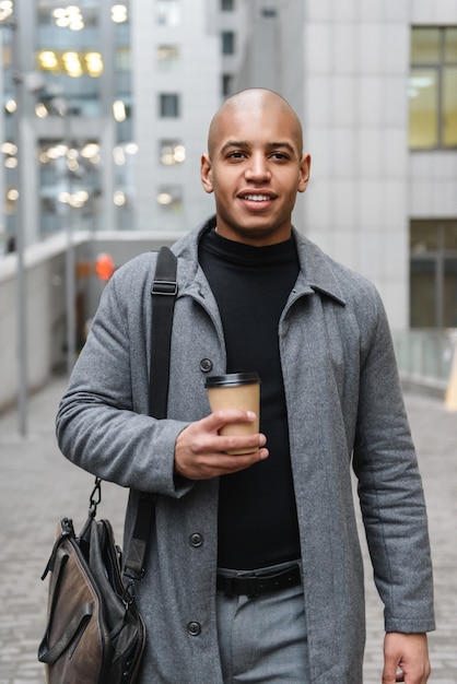 Aantrekkelijke jonge afrikaanse man die een herfstjas draagt terwijl hij in de straat van de stad staat en een koffiekopje drinkt