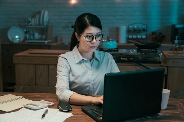 Aantrekkelijke jonge aziatische chinese vrouw die 's nachts in huiskeuken werkt. kantoordame met behulp van laptopcomputer op donkere werkplek om middernacht. geconcentreerde vrouwelijke werknemer in glazen met behulp van notebook pc.