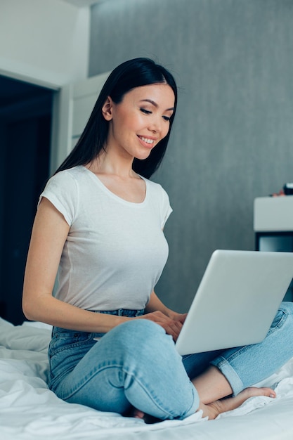 Aantrekkelijke jonge blanke vrouw zittend op een bed in jeans en wit T-shirt en met behulp van een moderne laptop