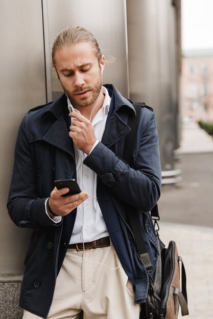 Aantrekkelijke jonge blonde man in formele kleding praten via oortelefoons, mobiele telefoon vasthouden terwijl hij buiten op een straat in de stad staat