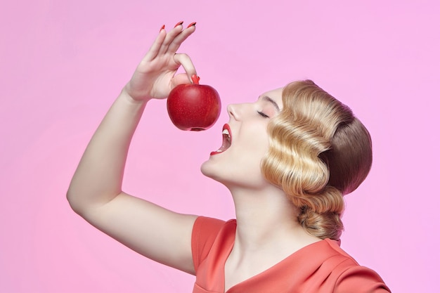 Aantrekkelijke jonge blonde met een retro kapsel staat op het punt in een rode appel te bijten