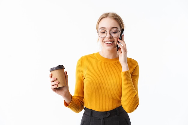 Aantrekkelijke jonge blonde vrouw die een trui draagt die over een witte muur staat, op een mobiele telefoon praat en een koffiekopje vasthoudt