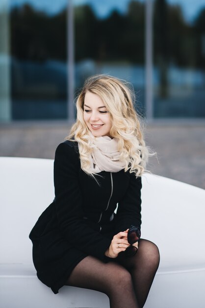 Aantrekkelijke jonge blonde vrouw met mooie make-up, het dragen van casual street wear, zittend op een witte plastic bank voor tegels muur van het gebouw