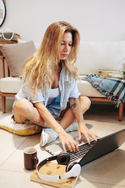 Aantrekkelijke jonge blonde vrouw zittend op de vloer in slaapzaal en huiswerk voor haar online klas