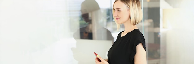 Aantrekkelijke jonge blonde zakenvrouw in stijlvolle zwarte blouse met smartphone in bedrijfskantoor