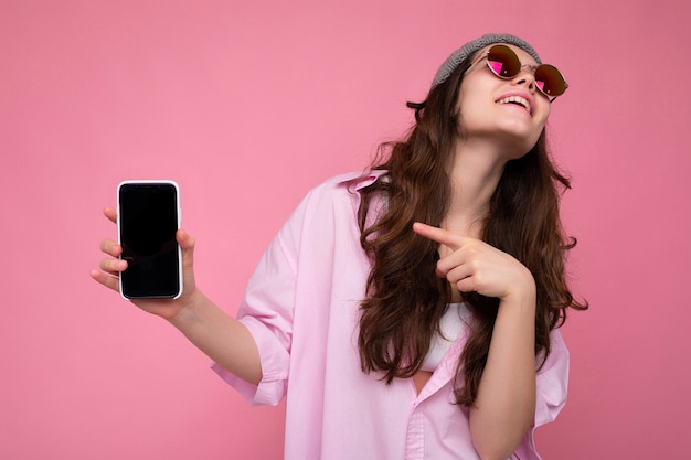 Aantrekkelijke jonge brunette vrouw met een stijlvolle roze shirt, grijze hoed en kleurrijke zonnebril