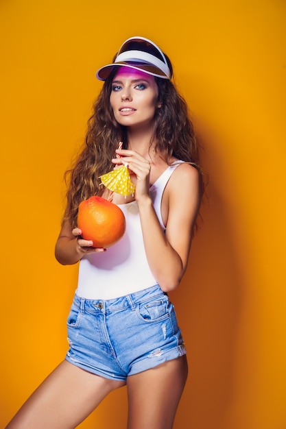 Aantrekkelijke jonge dame in zomeruitrusting die verse, sappige citrus met stro en paraplu houdt en camera bekijkt terwijl hij op geel staat