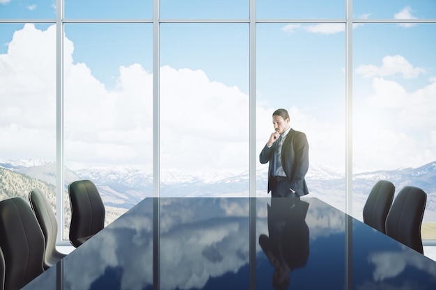 Aantrekkelijke jonge Europese man denken in moderne vergaderruimte interieur met reflecties op tafel stoelen en panoramisch raam met uitzicht op de wolken en daglicht Ceo en conferentie concept