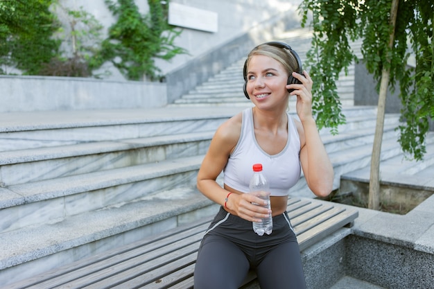 Aantrekkelijke jonge fitness blonde vrouw luistert naar muziek op een koptelefoon en houdt een fles water buiten. Gezond levensstijlconcept