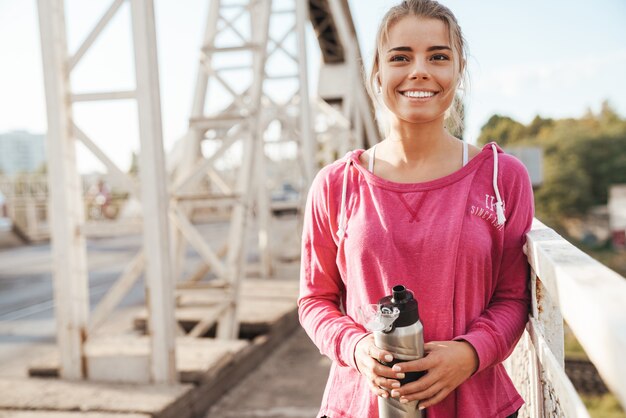 Aantrekkelijke jonge fitnessvrouw die buiten op de brug traint, naar muziek luistert met een koptelefoon, een waterfles vasthoudt