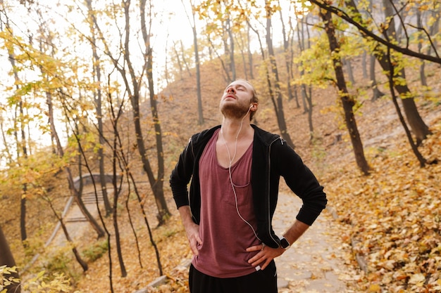 Aantrekkelijke jonge fitte sportman joggen in het herfstbos, luisteren naar muziek met oortelefoons, rusten