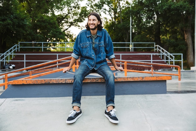 Aantrekkelijke jonge hipster man zit in het skatepark met een skateboard