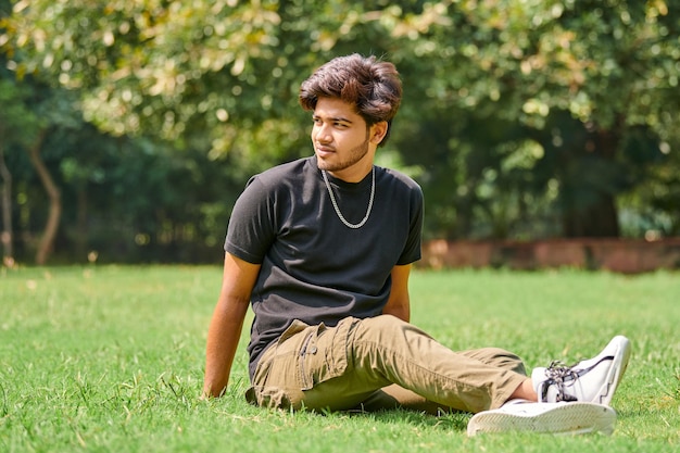 Aantrekkelijke jonge Indiase man portret in zwart t-shirt en zilveren nek ketting zittend op groen gazon