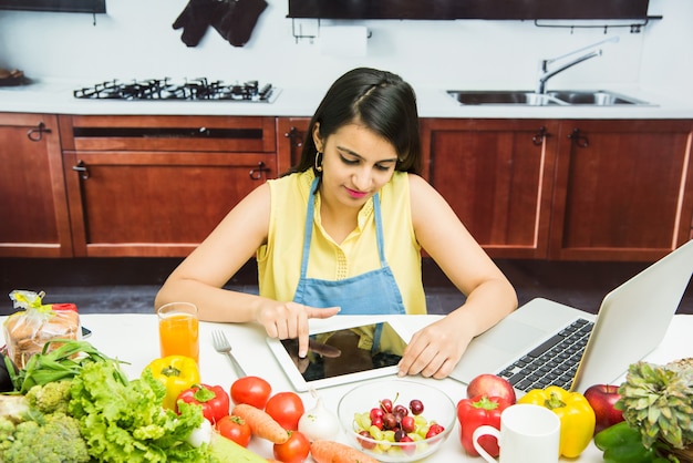 Aantrekkelijke jonge indiase meisje of vrouw met schort in keuken multitasking, met behulp van smartphone, tablet pc met tafel vol groenten en fruit en computer