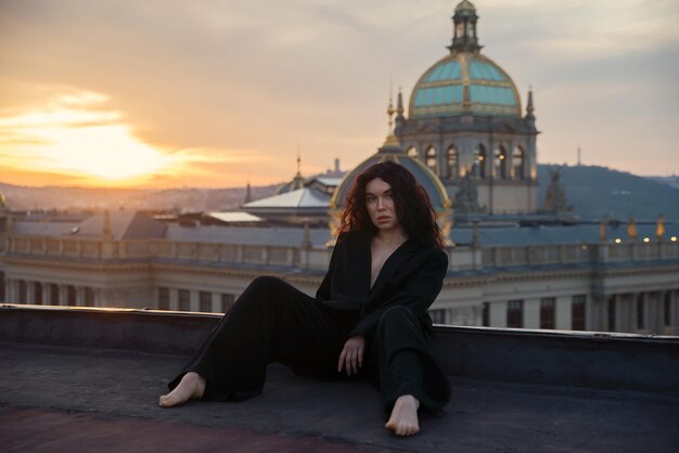 Foto aantrekkelijke jonge krullende brunette vrouw in een zwart jasje zonder lingerie zit op een dak tegen een