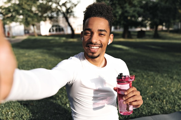 Aantrekkelijke jonge, krullende, donkere man in een wit sport-t-shirt glimlacht oprecht, houdt een roze waterfles vast en neemt een selfie in het park