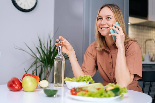 Aantrekkelijke jonge lachende vrouw praten aan de telefoon aan de tafel met groenten, fruit en salade in de keuken met modern interieur Concept van gezond eten