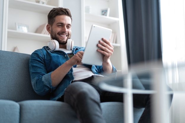 Aantrekkelijke jonge man gebruikt een tablet en glimlacht terwijl hij thuis op de bank ligt.