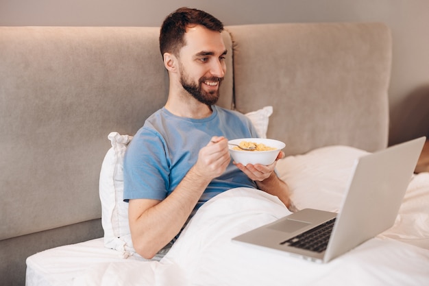 Aantrekkelijke jonge man met baard glimlachend eten ontbijt op bed voor laptop