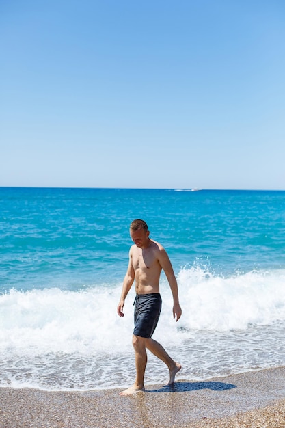 Aantrekkelijke jonge man met een mooi lichaam in korte broek loopt langs de zanderige kust en geniet van de zomervakantie