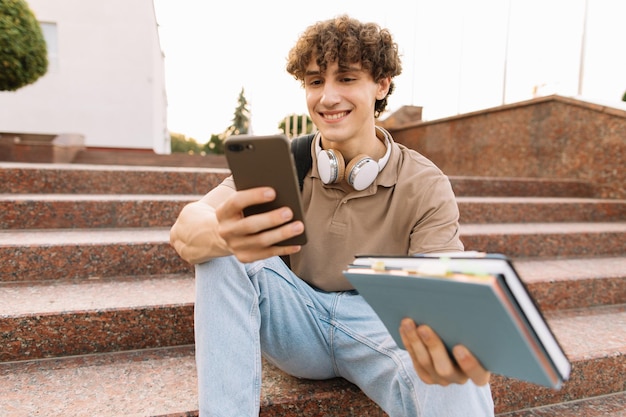 Aantrekkelijke jonge man met krullend haar, universiteits- of hogeschoolstudent zit op de trap en kijkt naar een smartphone