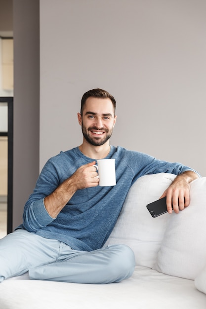 Foto aantrekkelijke jonge man ontspannen op een bank thuis, met behulp van mobiele telefoon, koffie drinken