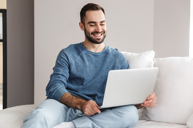 Aantrekkelijke jonge man ontspannen op een bank thuis, werken op laptopcomputer
