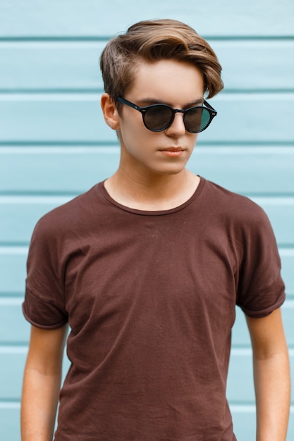 Aantrekkelijke jonge modieuze hipster man in zwarte stijlvolle zonnebril in trendy bruin t-shirt vormt in een stad in de buurt van een blauw houten gebouw op een zomerdag