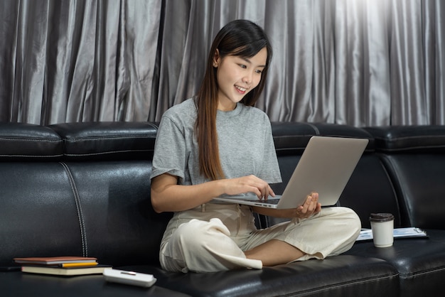Aantrekkelijke jonge mooie Aziatische vrouw die met laptop en document werken terwijl het zitten op het binnen woonkamerbureau als freelancer, e-Coachend, op afstand werkend of werk vanuit huisconcept.
