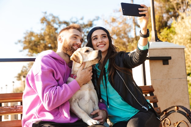 Aantrekkelijke jonge paar zittend op een bankje op het strand met hun hond, een selfie nemen