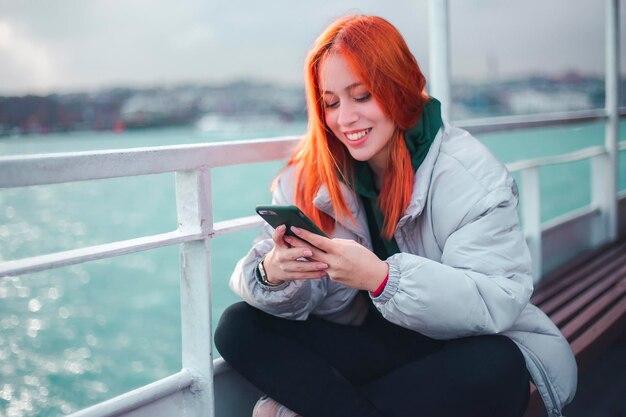 Aantrekkelijke jonge roodharige vrouw die een bericht typt via haar mobiele telefoon in een veerboot in Istanbul en ze glimlacht