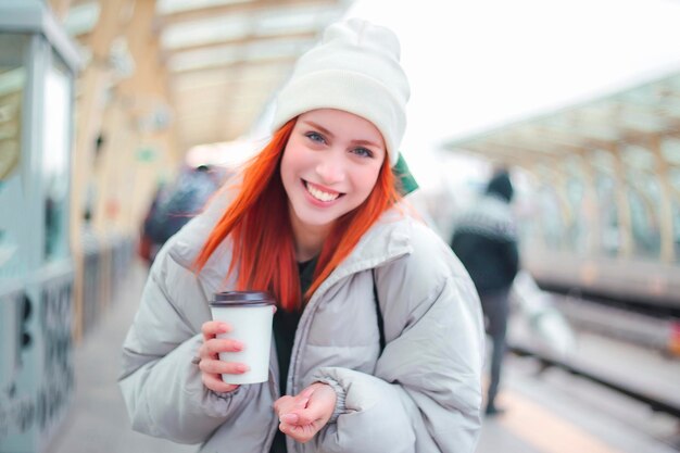 Aantrekkelijke jonge roodharige vrouw die lacht en naar de camera kijkt om een koffie mee te nemen in haar hand